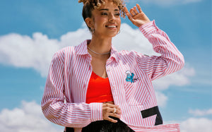 Female model shading her eyes with one hand against a cloudy blue sky, wearing a red and white striped button-up with an embroidered Smurf peeking from the chest pocket, open over a red bra.