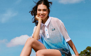Female model resting her chin on her hand under a blue sky, wearing a white-to-blue mesh polo with a white collar, light blue buttons, and a 'ZUMBA' X 'THE SMURFS' chest patch.