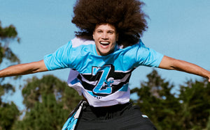 Male model leaping with arms spread against trees and blue sky, wearing a light blue, black, and white jersey with a large 'Z' and a Smurf, over black pants.
