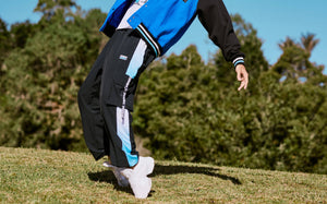 Cropped view of a male model leaning mid-dance on a grassy hill, wearing black cargo pants with light blue mesh side panels and a 'ZUMBA' webbing strap under a blue and black varsity jacket, with trees behind him.