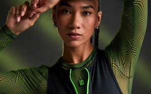 Close-up of a female model with one arm raised over her head, wearing a neon green and black wavy check print mesh long sleeve top with a small 'STRONG iD' patch under a black bodysuit with a neon green zipper. Dark background with a soft green glow.