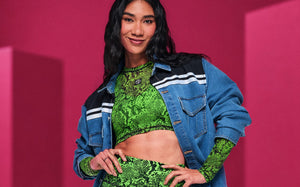Female model with hands on hips in a green snakeskin-print long-sleeve crop top and matching biker shorts, under an open denim shacket with black and white striped shoulders, against a pink backdrop.