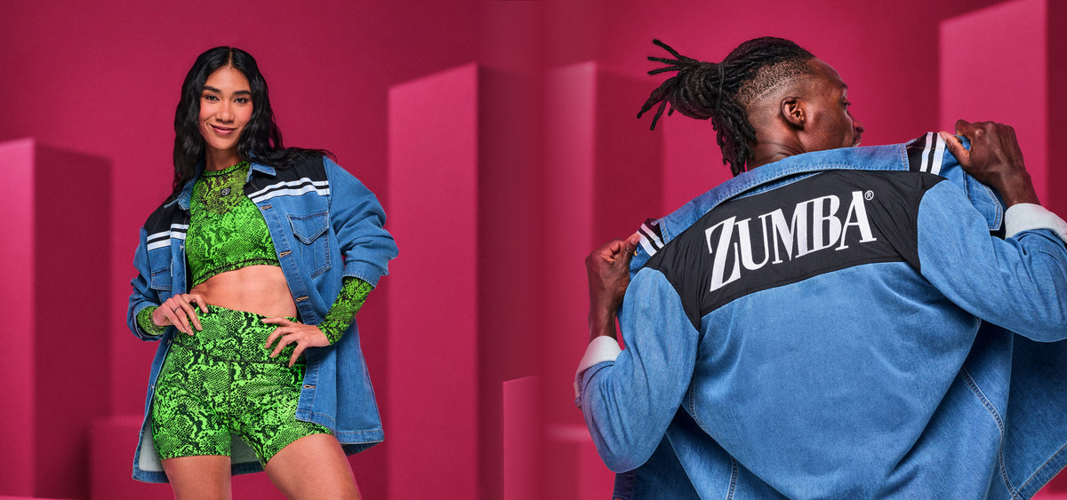 Two models against a pink backdrop with block shapes: on the left, a female model in a green snakeskin-print crop top and biker shorts under an open denim shacket with striped shoulders; on the right, a male model shown from the back holds open a denim shacket with 'ZUMBA' text in white at the shoulders.