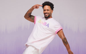 Male model in a white Zumba logo T-shirt, smiling and posing against a soft purple gradient background.