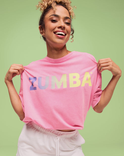 Smiling model holding a pink Zumba logo T-shirt at the shoulders with gradient lettering, paired with white drawstring shorts on a light green backdrop.