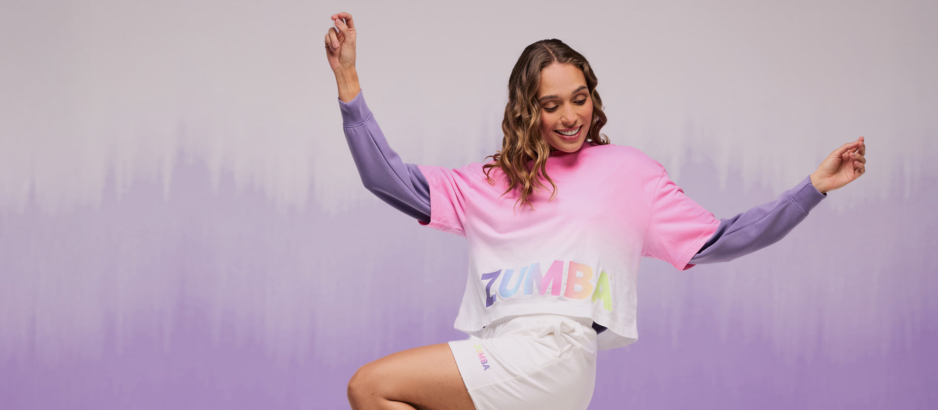 Smiling female model wearing a pink-to-white ombré Zumba cropped crop top with rainbow logo styling, layered over long purple sleeves of a sweatshirt and matched with white shorts. 