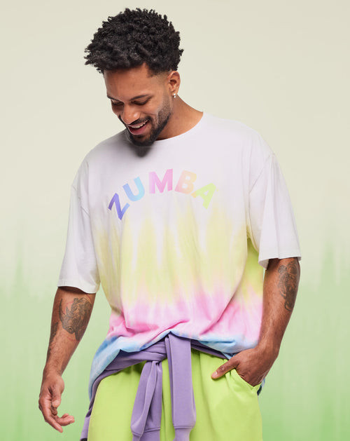 Male model wearing a white oversized Zumba logo T-shirt with pastel gradient fade and rainbow lettering with a lavender sweatshirt tied around his waist and lime green cargo pants against a soft green studio backdrop.