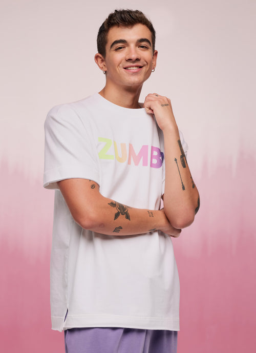 Smiling model wearing a white Zumba logo T-shirt with rainbow lettering, paired with lavender pants on a soft pink gradient backdrop.