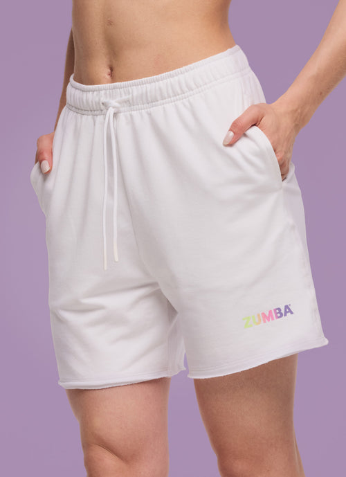 Close-up of white Zumba drawstring shorts with side pockets and small multicolor logo, shown on model against a lavender backdrop.