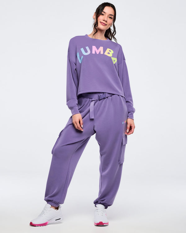 Purple “ZUMBA” cropped sweatshirt with multicolor gradient lettering, relaxed fit, and crewneck, worn by a female model with matching purple cargo joggers, belt detail, and white sneakers against a light studio background.