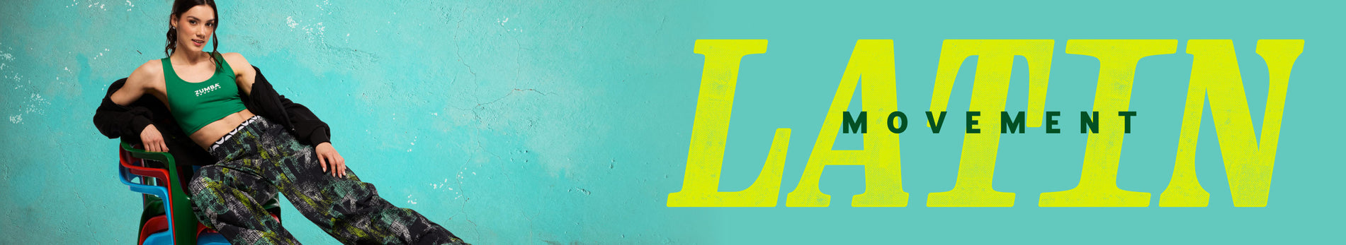 Banner with a turquoise textured background, a female model reclining on stacked colorful chairs at left, and bold yellow “LATIN” text with smaller “MOVEMENT” across the center. Featuring Zumba dance wear: she wears a green Zumba sports bra, a black hoodie draped off her shoulders, loose black wide-leg pants with an abstract green-and-gray print and a branded waistband, and neon green lace-up sneakers.