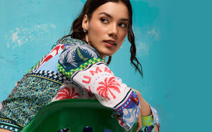Close-up lifestyle shot of a female model leaning on a green chair against a teal wall, wearing a multi-colored shirt.