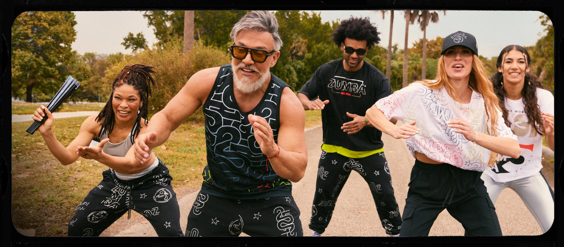 Banner showing five models dancing together on a tree-lined road outdoors. The product features Zumba activewear on male and female models.