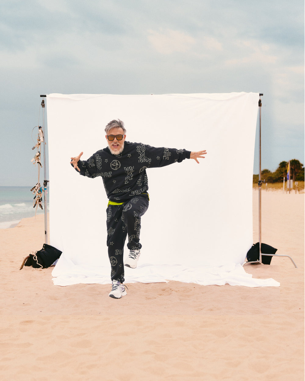 Black Zumba sweatshirt and jogger set with silver allover logo print. Male model and Zumba founder Beto wears the relaxed long-sleeve sweatshirt, matching joggers and white sneakers, while posing on a sandy beach in front of a white backdrop.