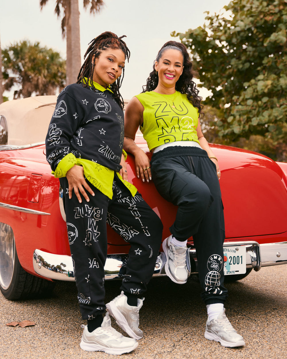 2 female models leaning against a vintage red car. The model on the left wears a black Zumba sweatshirt with silver allover logo print, matching black joggers, and silver sneakers. Female model on the right wears a neon yellow sleeveless Zumba crop top, black joggers, and silver-and-white sneakers.