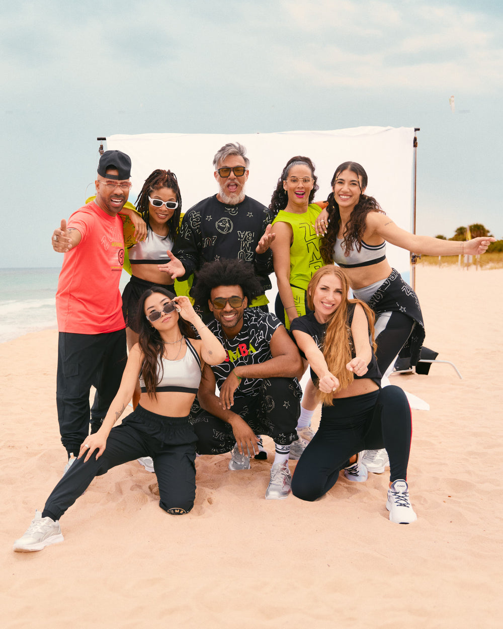 Group lifestyle photo on a beach featuring Zumba apparel. Eight male and female models pose together in branded activewear, including graphic tops, sports bras, black joggers, leggings, biker shorts, sneakers, neon coral T-shirt and a black hat.