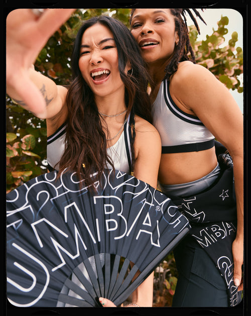 Black folding fan with oversized silver Zumba lettering. Two female models wear silver sports bras and black logo-print bottoms, posing closely outdoors with the fan in the foreground.
