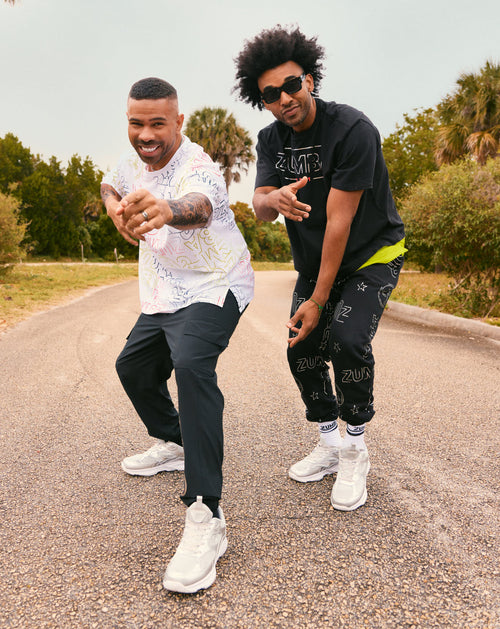 Two men who model Zumba streetwear outdoors on a road. Male model on the left wears a white short-sleeve button-up shirt with pastel allover print, black pants, and silver sneakers; male model on the right wears a black Zumba T-shirt, black logo-print joggers, white socks and silver sneakers.