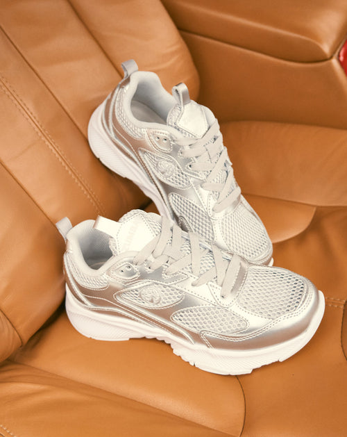 White and metallic silver Zumba sneakers with mesh panels, lace-up closure, and chunky white soles. The pair is displayed on a tan leather car seat.