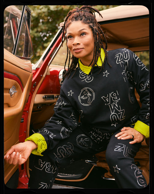 Black Zumba sweatshirt and jogger set with silver allover logo print and a neon yellow collar and cuff accents of the button-down underneath. Female model wears this while sitting in a red car with a tan, leather interior.