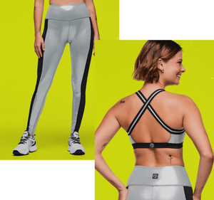 Silver high-waisted leggings with black side panels shown with a matching silver sports bra featuring black trim and crisscross back straps. Female model wears this against a neon yellow background.