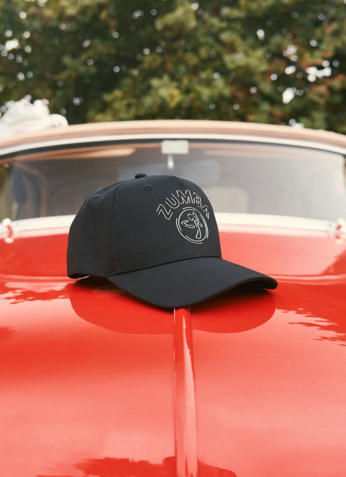 Product close-up highlighting a black Zumba baseball cap with a silver Zumba logo sits on the hood of a shiny red vintage car, with the windshield and blurred green trees in the background.
