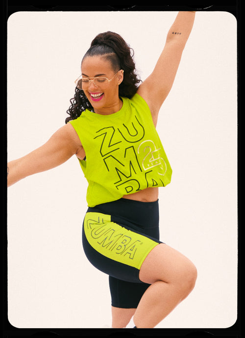 A female model smiles in a neon green cropped sleeveless Zumba top paired with high-waisted black-and-neon green and bike shorts modeling in front of a white background.