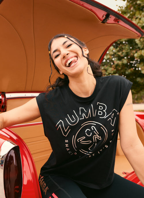 A female model sits at the open trunk of a red vintage car wearing a black sleeveless Zumba graphic top and black leggings with a side logo detail.