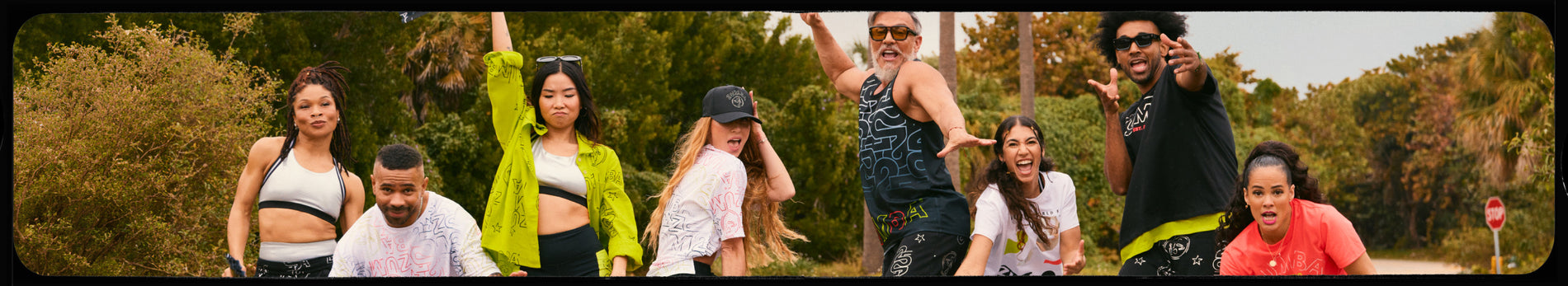 Banner showing a lively outdoor group dance scene on a road lined with trees and palm trees. The image highlights Zumba apparel on male and female Zumba Educational Specialists, posing energetically in coordinated workout outfits, with Beto in the middle. 
