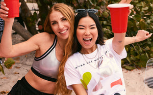 Beach party-style photo featuring Zumba activewear. Two female models smile and raise red cups. One wears a metallic silver sports bra with black trim and black shorts; the other wears a white graphic T-shirt with colorful print.
