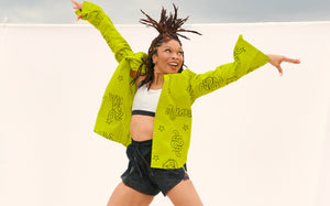 A female model poses in a neon green oversized button-down Zumba shirt over a silver sports bra, paired with loose black shorts against a white backdrop.