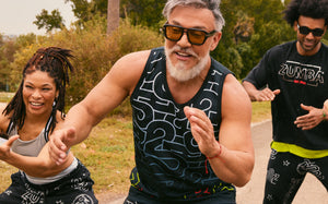 Outdoor action photo featuring men’s and women’s Zumba apparel. Beto, the founder of Zumba in front wears a black graphic tank and matching black shorts, while another male model behind wears a black Zumba T-shirt with black printed joggers. A female model at left wears a silver sports bra and black bottoms.