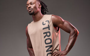 Banner featuring a male model in a beige sleeveless training top against a gray studio background. The top has a clean athletic cut with oversized arm openings and bold vertical black “STRONG” lettering down the front. He stands with hands on hips, highlighting the relaxed fit and sporty design.