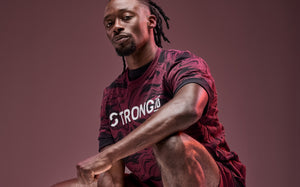 Banner featuring a male model in burgundy performance activewear against a mauve studio background. He wears a burgundy and black marbled short-sleeve athletic T-shirt with “STRONG iD” printed across the chest, paired with matching burgundy shorts. The squatting pose highlights the sporty coordinated set and the bold graphic pattern.