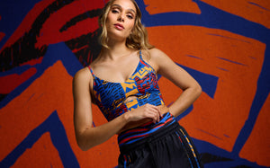 Female model wears a blue, red, and yellow printed crop top with a front cutout and black pants with a matching printed waistband. She stands in front of a vivid orange and blue abstract mural.