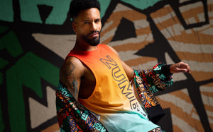 Male model wears an orange-to-mint gradient Zumba tank with black logo lettering and a multicolor geometric print jacket draped off the shoulders. He stands in front of a bold black, green, tan, and cream abstract mural.
