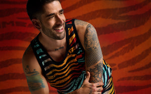 Male model wears a black sleeveless tank with multicolor zebra stripes and matching shorts. He poses in front of a red and orange zebra-inspired backdrop.