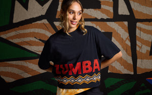 Female model wears a black cropped Zumba T-shirt with red lettering and geometric trim, layered over a yellow printed top. She stands in front of a bold black, tan, cream, and green abstract mural.
