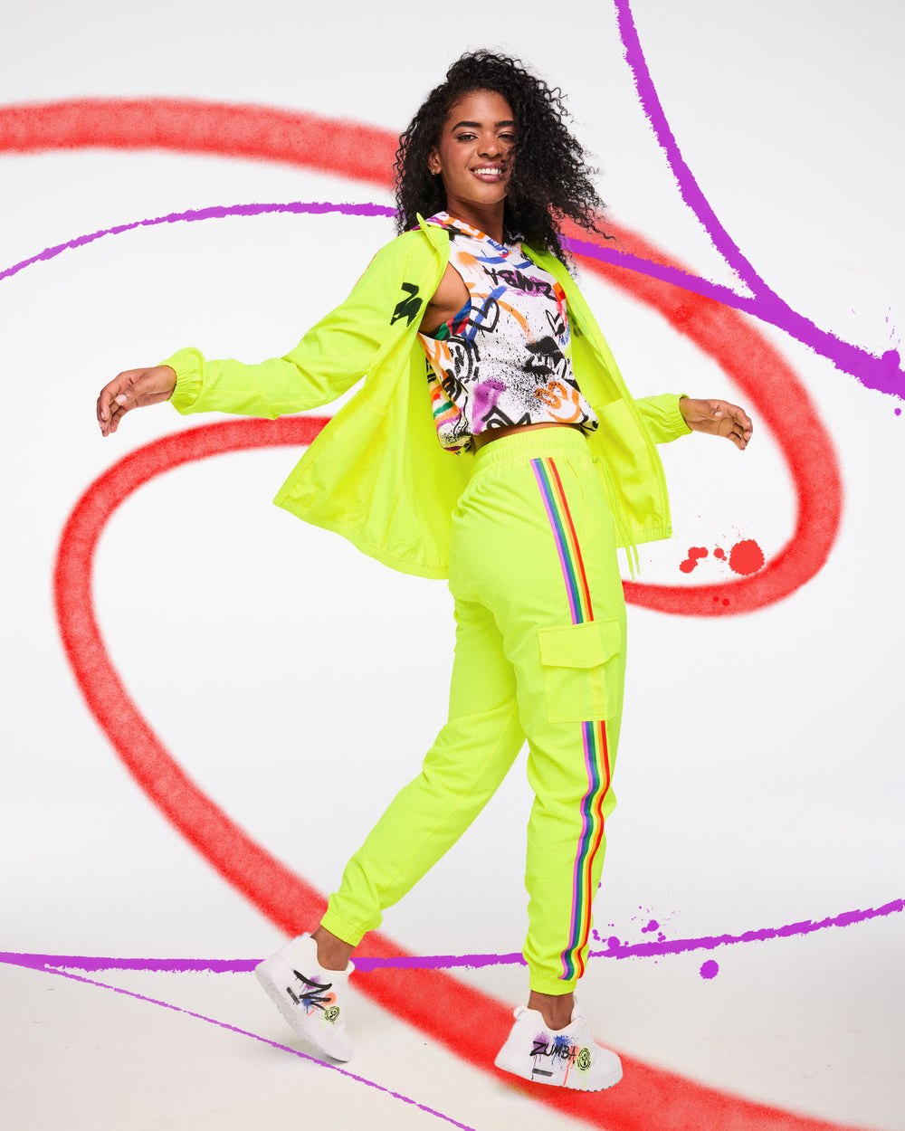 Woman in neon yellow Zumba outfit with rainbow‑stripe joggers dancing against a colorful spray‑paint backdrop.