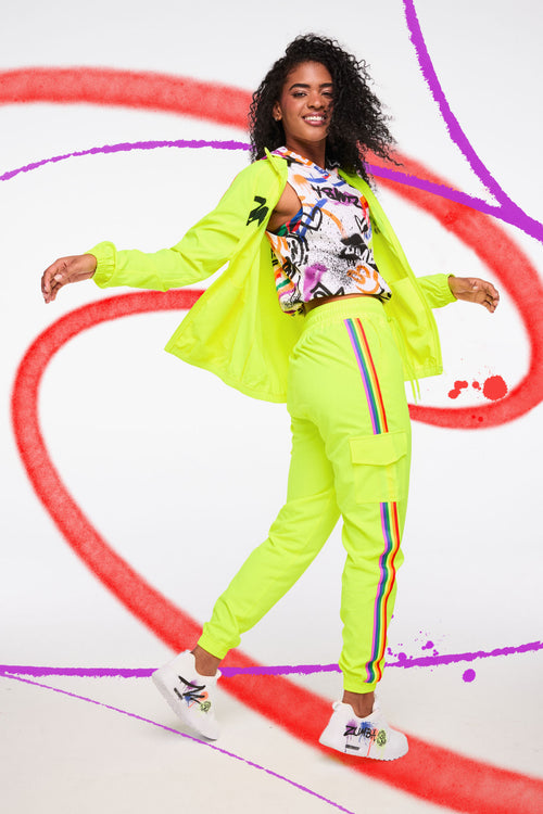 Female model in neon yellow jacket and rainbow‑stripe joggers with a graffiti crop top on a bold paint‑stroke background.
