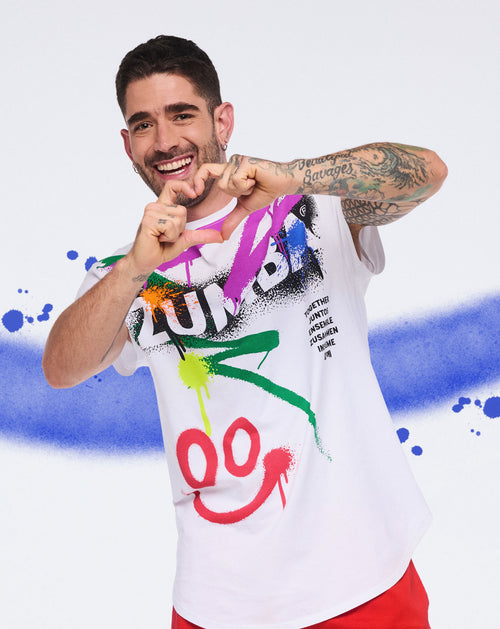 Smiling man in a white Zumba graffiti‑style shirt forming a heart shape against a colorful paint‑splash background.