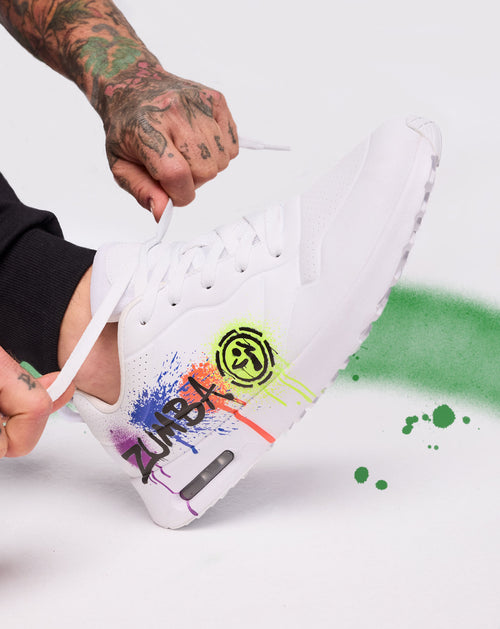 White Zumba sneaker with rainbow paint splashes as a tattooed hand ties the laces on a white backdrop with green spray.