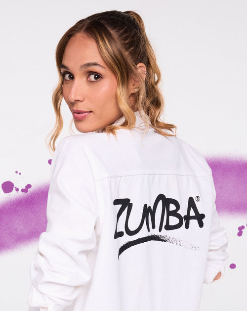 Woman in white Zumba shirt looking over shoulder against a white backdrop with purple paint splashes.