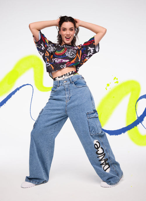 Female model in a black graffiti‑print crop top and wide‑leg denim cargo pants with “DANCE” graphic, set against bright neon paint strokes.
