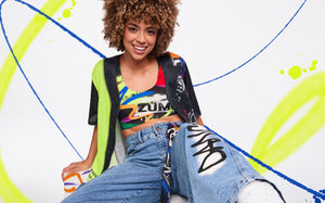 Female model in an open black graffiti‑print mesh button‑down over a colorful Zumba bra, reclining upright on the floor against a white backdrop with neon paint strokes.