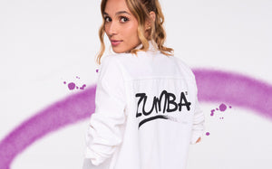 Woman in white shirt with bold “Zumba” logo poses against a clean backdrop with a purple spray‑paint swoosh.
