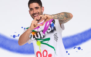 Smiling man in a colorful Zumba graphic tee forms a heart shape against a white backdrop with a blue paint swoosh.