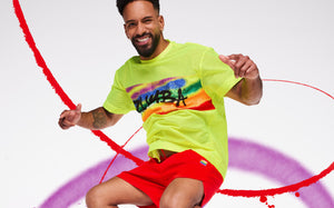 Smiling man in a neon yellow Zumba mesh tee and red shorts poses against a white backdrop with red and purple paint swirls.