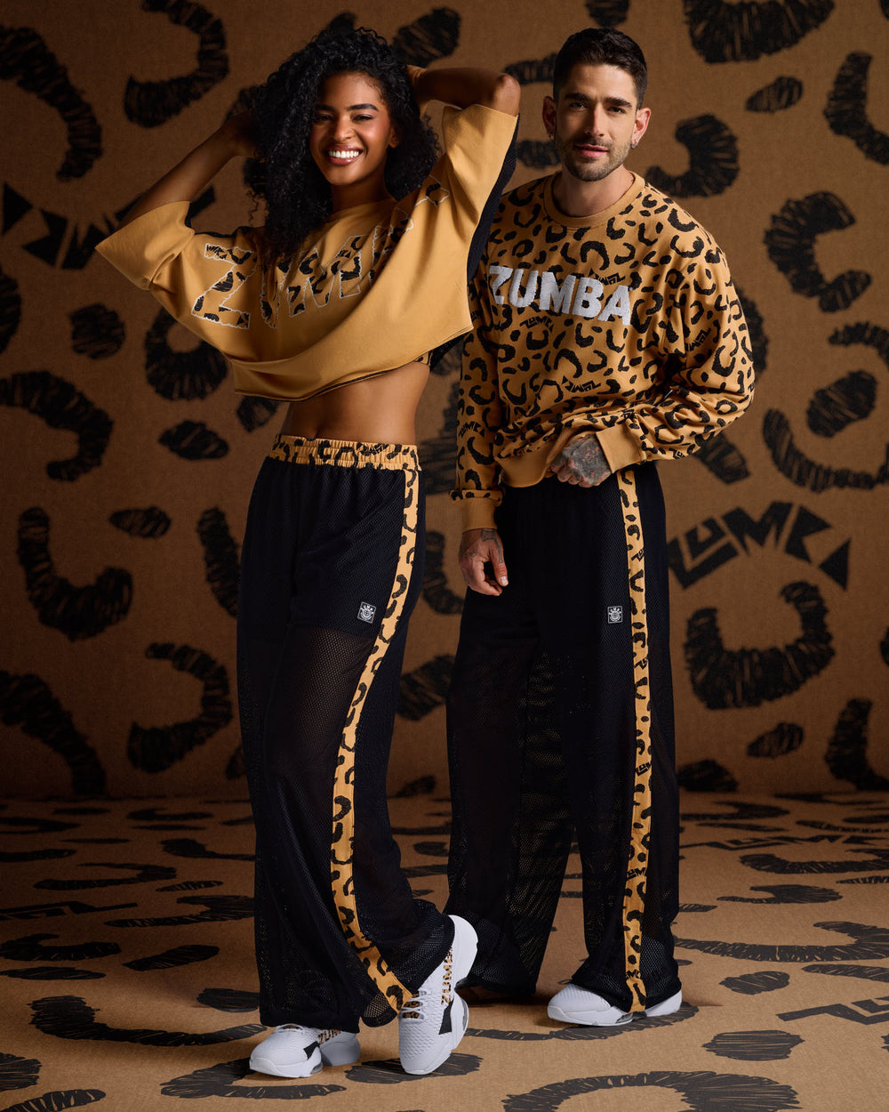 Male and female models wearing tan Zumba tops with cheetah print details and black mesh pants with cheetah trim, against a matching cheetah print background.