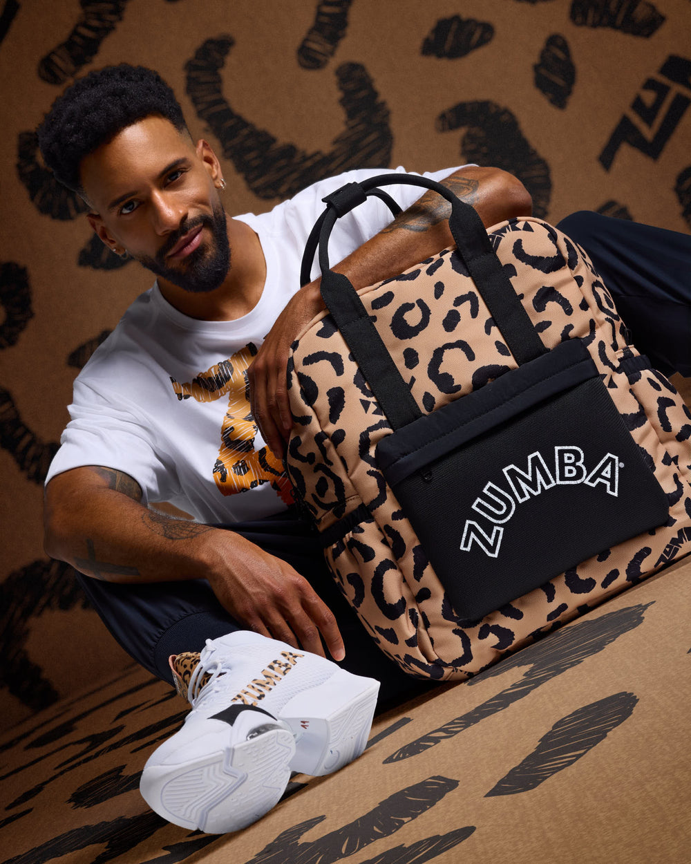 Male model wearing a white Zumba T-shirt and white sneakers, posing with a tan cheetah print Zumba backpack against a matching cheetah print background.